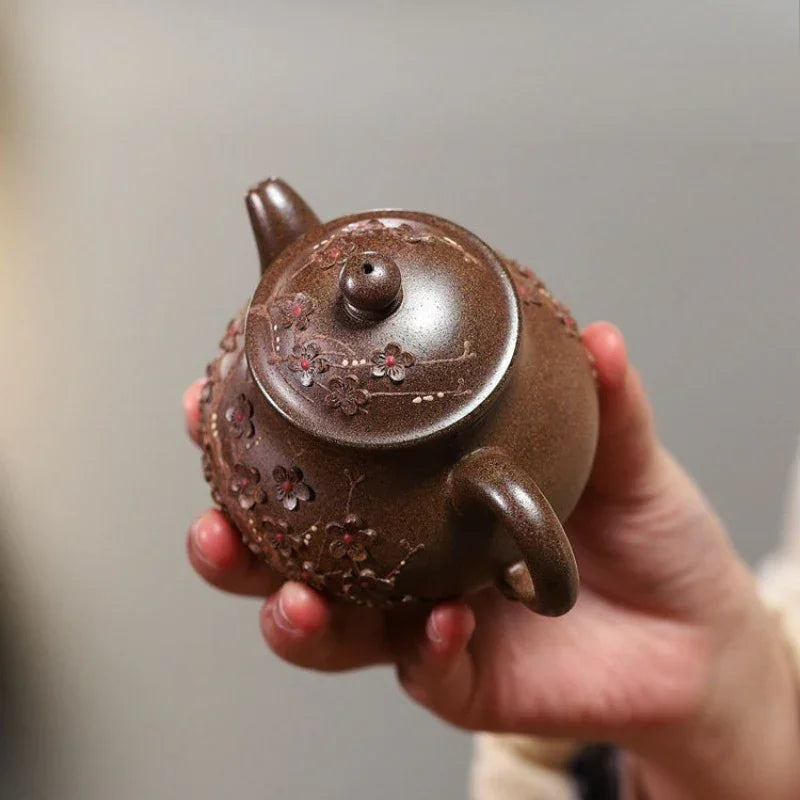 A hand holding a small brown ceramic teapot decorated with floral relief patterns, set against a soft, blurred gray background.

