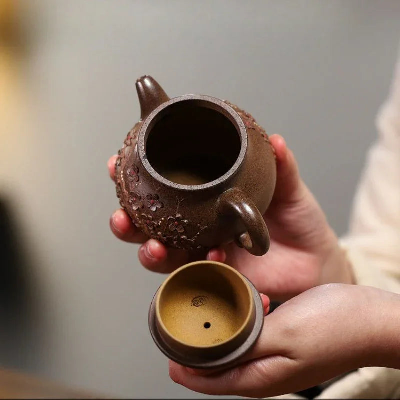 A person holds a small brown ceramic teapot with a matching lid in both hands. The teapot has a rounded body with textured floral details, and the interior is shown alongside the lid.

