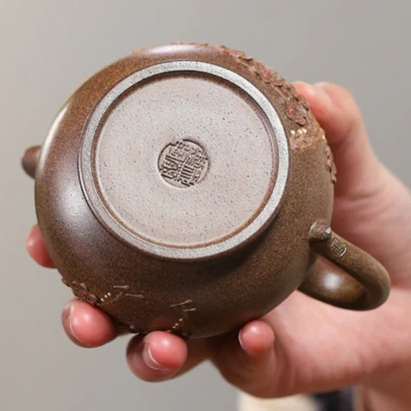 A close-up of a hand holding the base of a brown ceramic tea pot, showing a circular stamped seal on the bottom and textured clay details.

