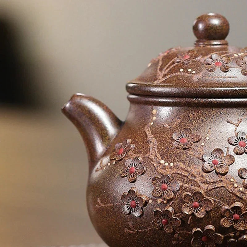 Close-up of a brown ceramic teapot decorated with raised cherry blossom flowers and branches, shown against a softly blurred neutral background.

