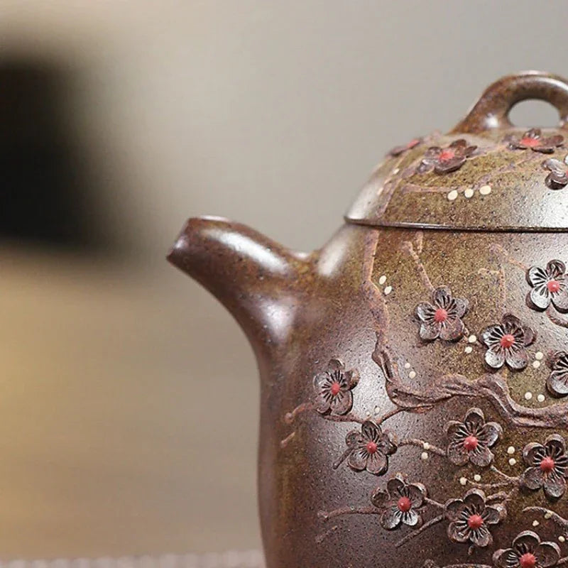 Close-up of a brown ceramic teapot decorated with raised cherry blossom flowers and small red centers, shown against a softly blurred neutral background.

