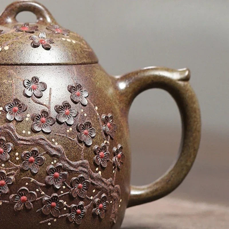 Close-up of a brown ceramic teapot decorated with raised cherry blossom branches and small red-pink flowers, shown against a soft gray background.

