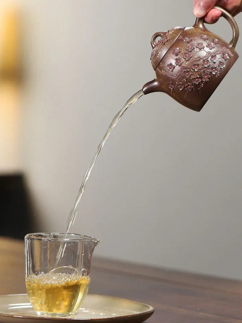 A hand holds a brown ceramic teapot with floral embossing, pouring tea from a clear glass pitcher on a plate into a cup.

