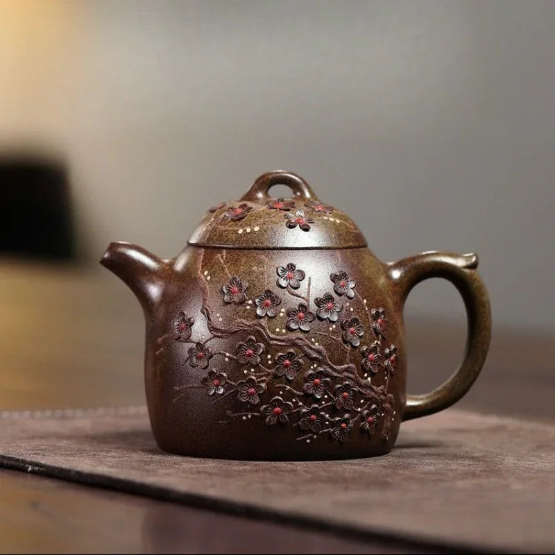 A dark brown ceramic teapot with embossed cherry blossom branches and red flower details, resting on a textured mat against a blurred gray background.

