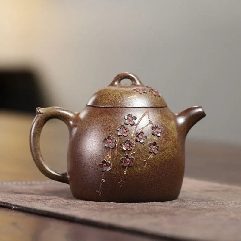 A brown ceramic teapot with a metallic bronze glaze and delicate cherry blossom designs, resting on a textured brown mat against a blurred neutral background.

