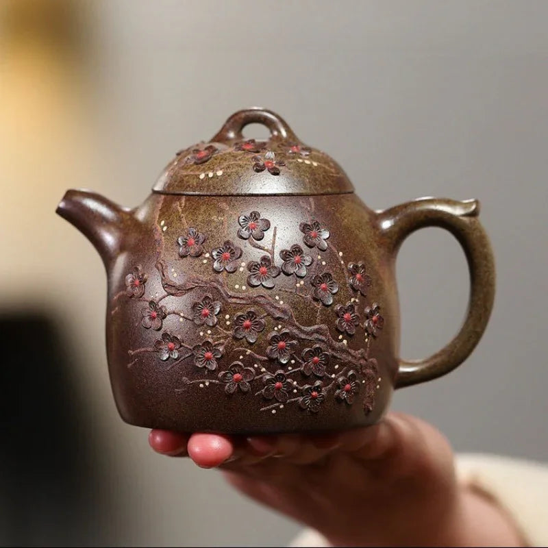 A hand holds a brown ceramic teapot decorated with raised floral patterns and small red accents, against a softly blurred gray background.

