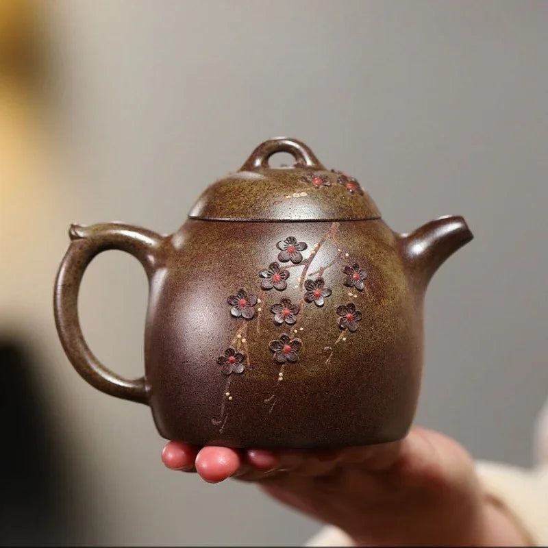 A hand holds a small brown ceramic teapot decorated with red and pink cherry blossoms, against a softly blurred gray background.

