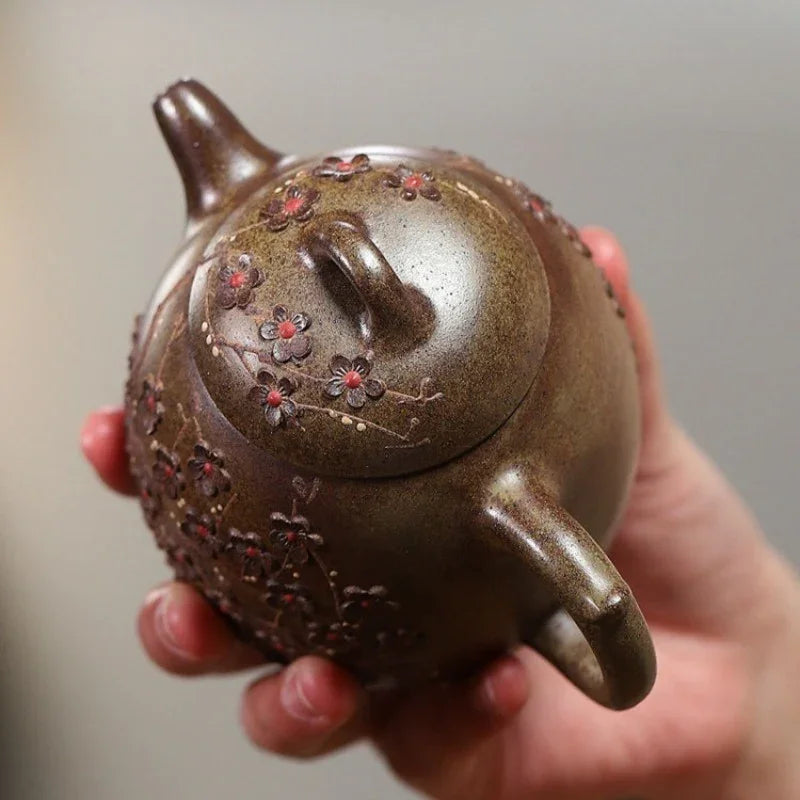 A hand holding a small brown ceramic teapot decorated with red and gold floral patterns, shown against a soft gray background.

