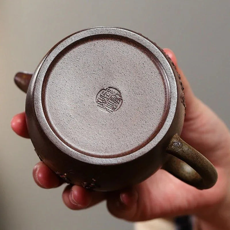 A close-up of a hand holding the base of a dark brown ceramic teapot, showing a circular stamped seal on the bottom and the side handle.

