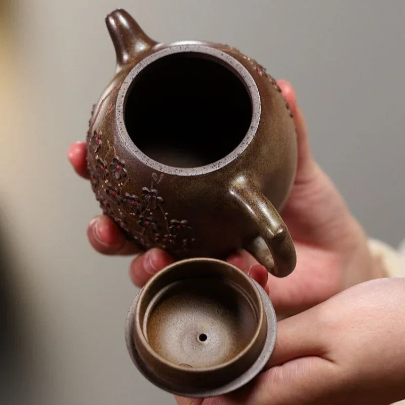 A close-up of a hand holding a brown ceramic teapot with a matching lid removed, showing the interior and textured exterior against a soft gray background.

