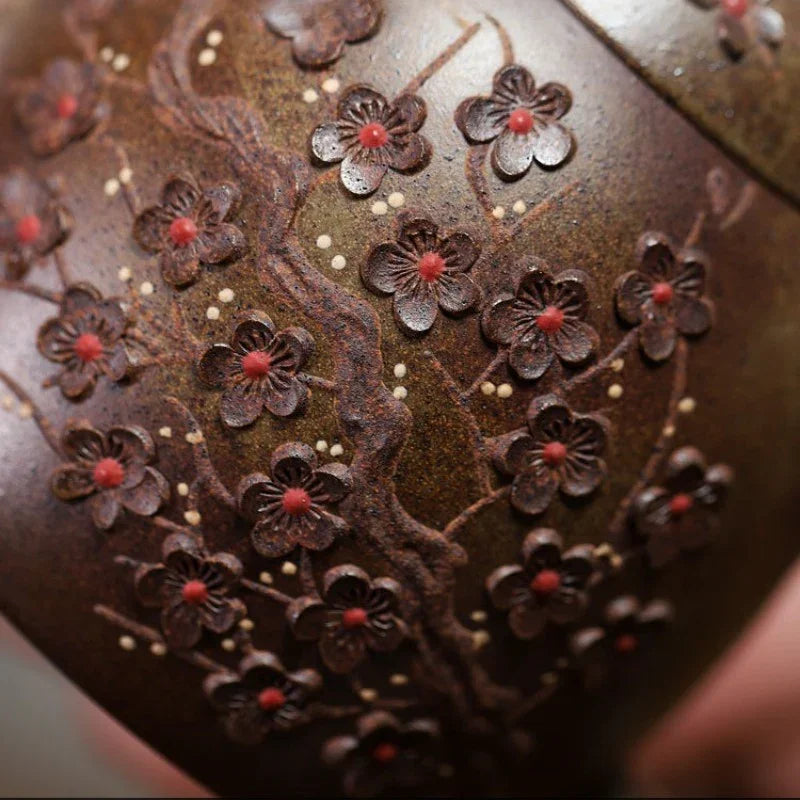 Close-up of a dark brown ceramic vessel decorated with raised cherry blossom motifs, each with red centers and small white dots, with a finger partially visible at the bottom right.

