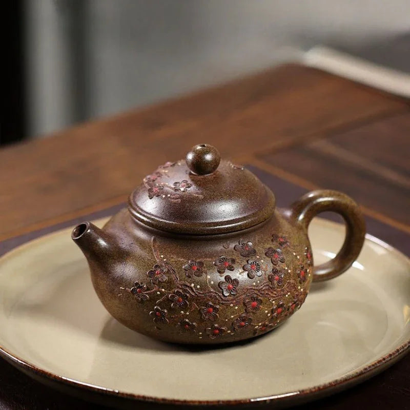 A brown ceramic teapot with floral relief details and red accents, resting on a beige ceramic plate over a dark wooden table.

