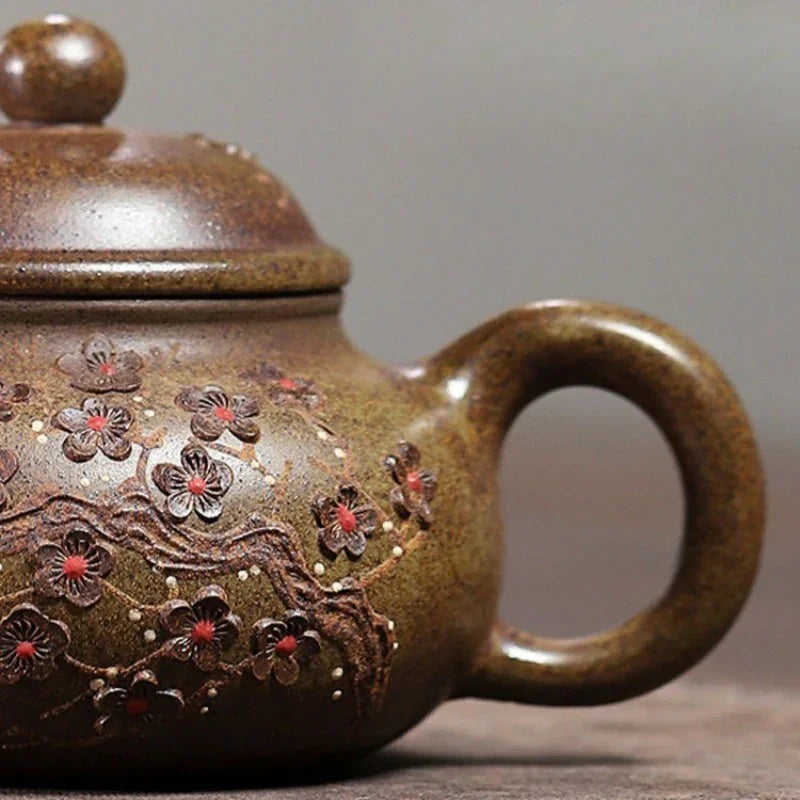 Close-up of a brown ceramic teapot with a rounded handle and lid, decorated with raised floral patterns in dark brown and red with small metallic accents, set against a neutral gray background.

