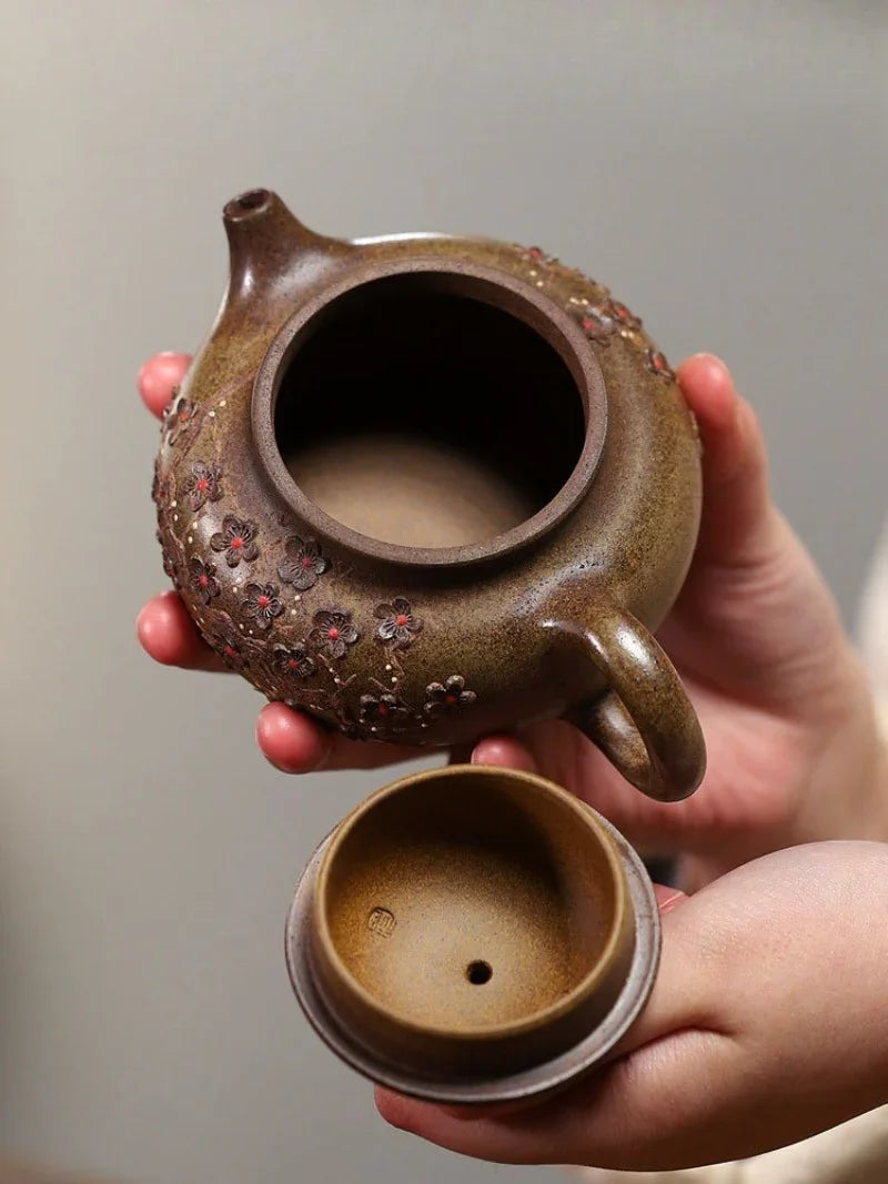 A close-up of a hand holding a brown ceramic teapot with a matching lid, featuring textured floral details on the side and a warm earthy glaze.

