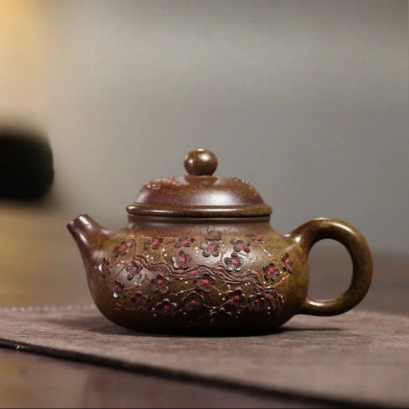 A brown ceramic teapot with floral relief patterns and red accents, resting on a dark wooden surface against a blurred gray background.

