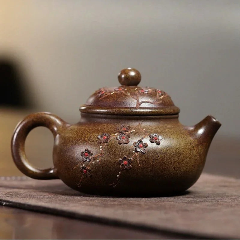 A brown ceramic teapot with a glossy glaze and hand-painted red and pink floral details, resting on a dark textured mat against a blurred gray background.

