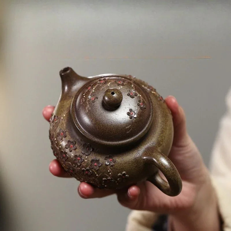 A hand holding a small brown ceramic teapot decorated with red and gold floral patterns, set against a soft gray background.

