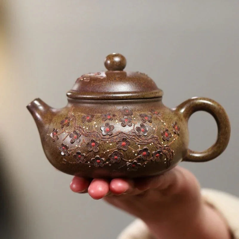 A small brown ceramic teapot with floral relief decorations and red accents, held in a person's hand against a soft gray background.

