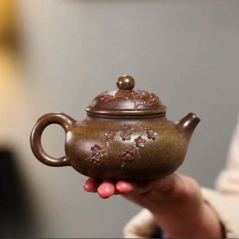 A small brown ceramic teapot with floral relief decorations, held in a person's hand against a softly blurred gray background.

