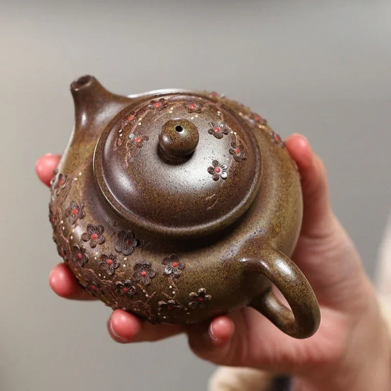 A hand holding a small brown ceramic teapot decorated with red and pink floral patterns, shown against a soft gray background.

