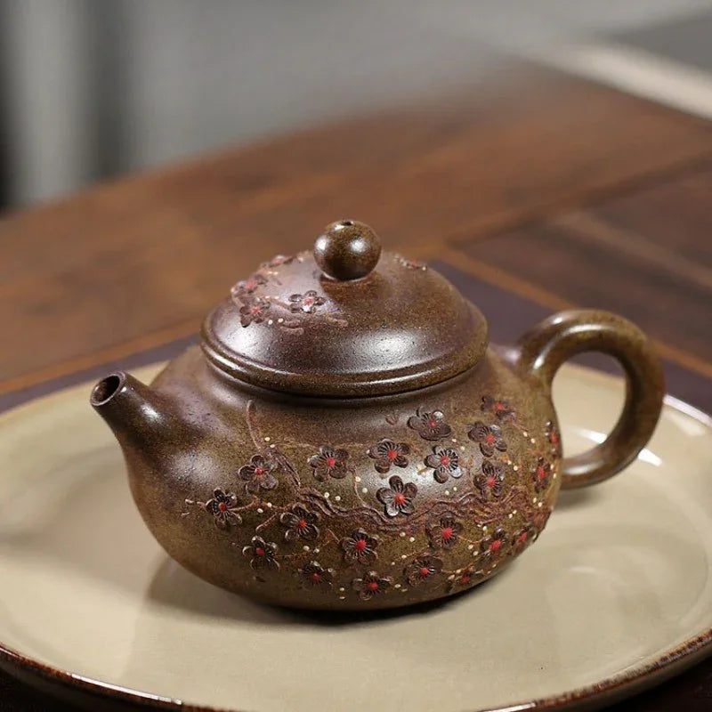 A brown ceramic teapot with red and gold floral embossed patterns, resting on a beige ceramic plate over a dark wooden table.

