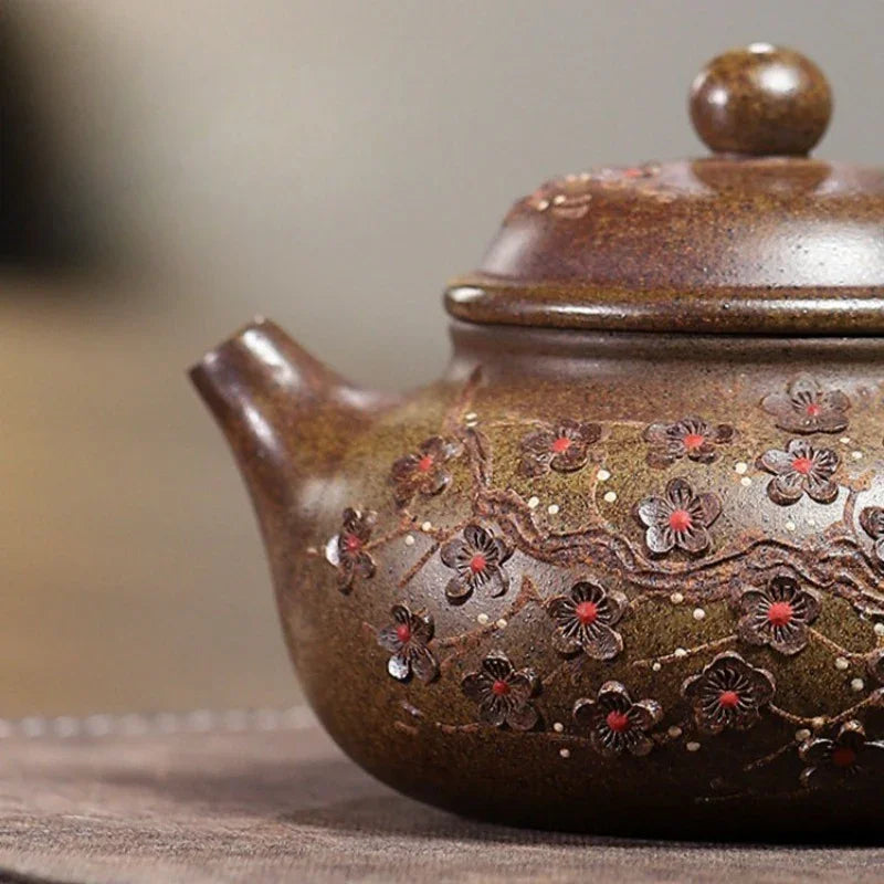 Close-up of a brown ceramic teapot with a glossy finish, decorated with raised floral motifs in dark brown and red, on a neutral background.

