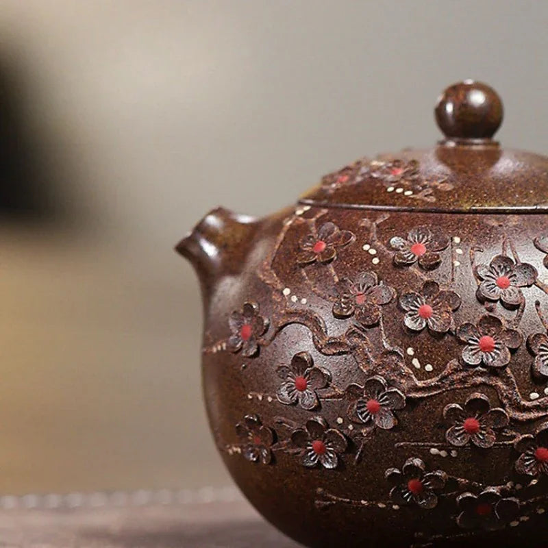 Close-up of a dark brown ceramic teapot decorated with raised floral motifs and small red centers, shown against a softly blurred neutral background.

