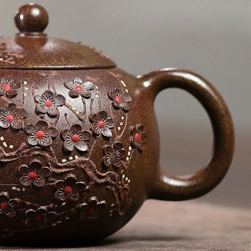 A close-up of a dark brown ceramic teapot decorated with raised floral patterns and small red accents, shown against a soft gray background.

