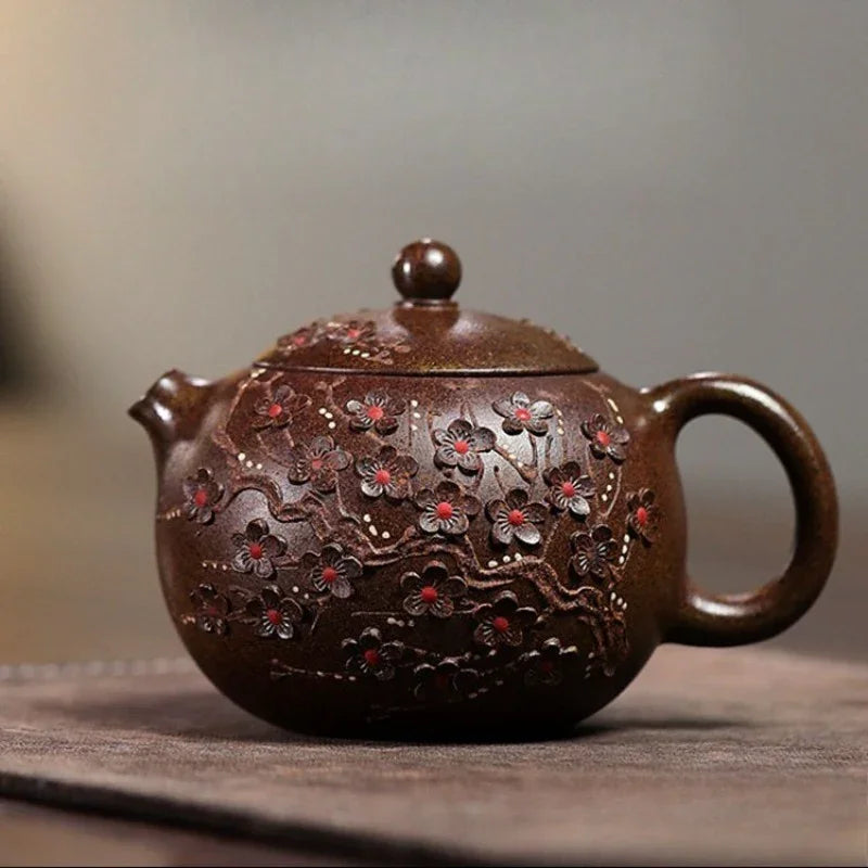 A dark brown ceramic teapot with hand-painted cherry blossom details, resting on a textured surface against a soft, blurred gray background.

