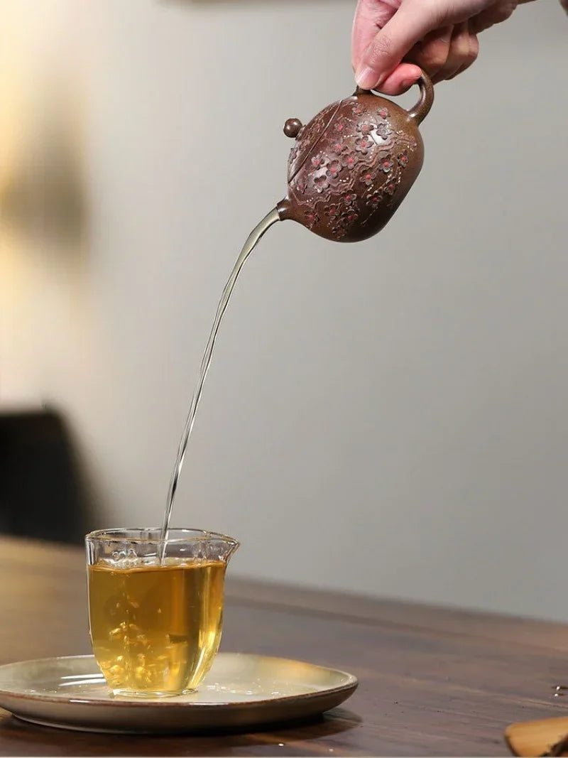 A hand holds a small textured brown teapot pouring tea into a clear glass cup on a saucer, set on a wooden table against a softly blurred background.

