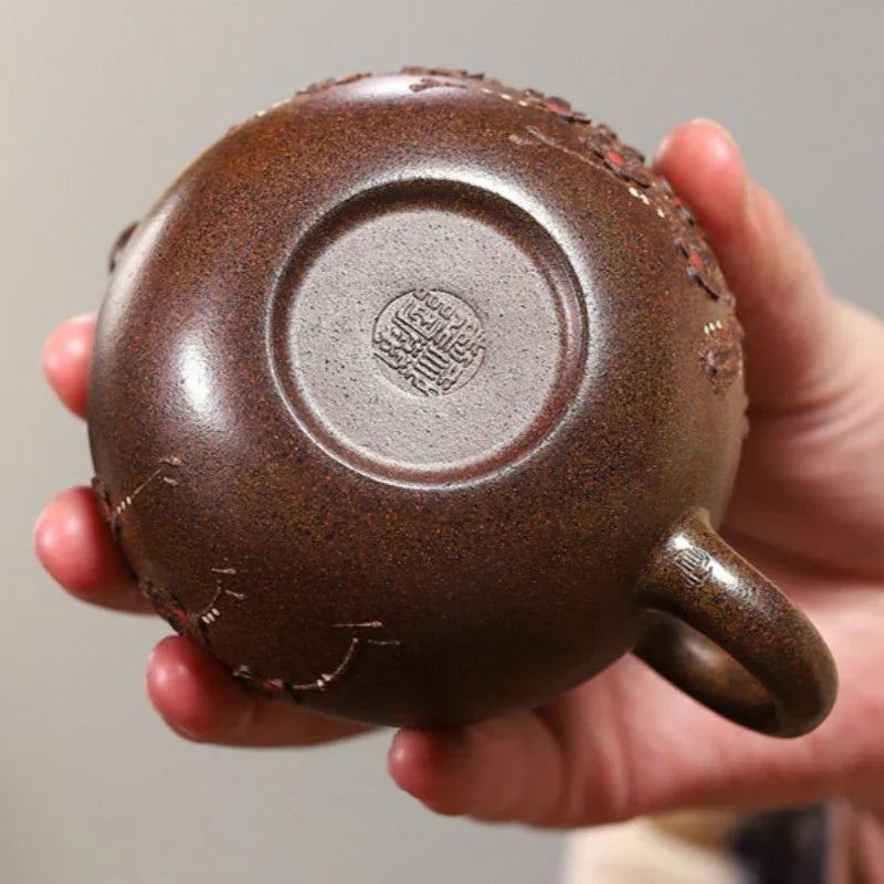 A close-up of a hand holding the base of a dark brown ceramic teapot, showing a circular stamped seal and the textured clay surface.

