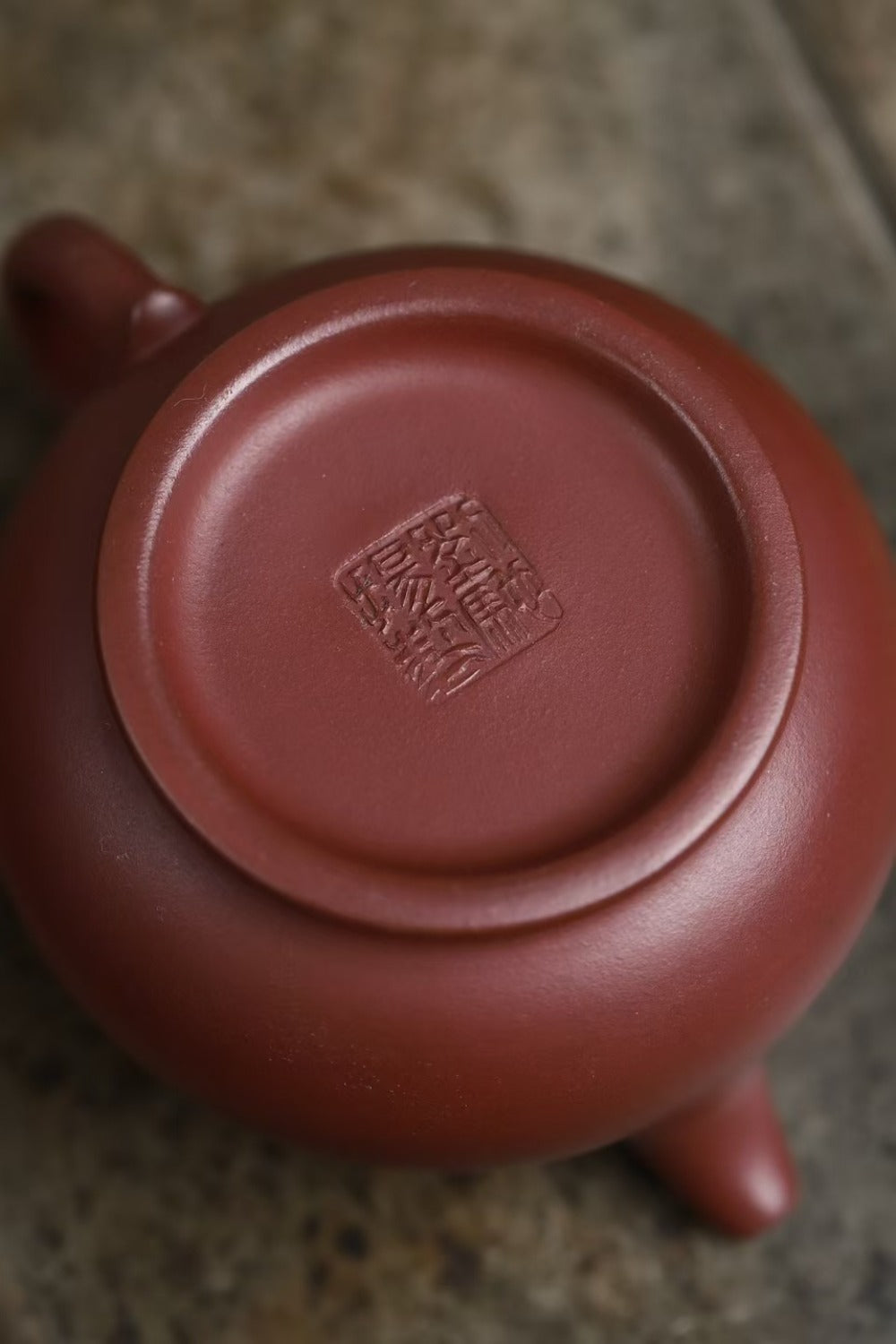 Close-up of the bottom of a reddish-brown clay teapot, showing a stamped seal mark in the center, resting on a textured stone surface.

