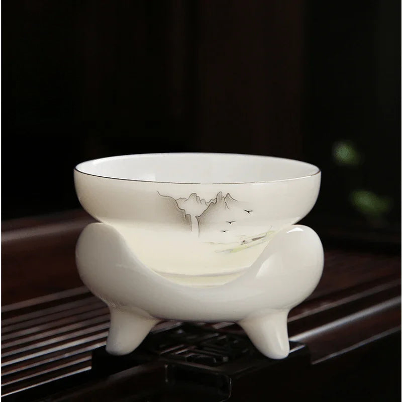A white ceramic tea bowl with a matching footed stand, decorated with delicate black and green landscape brushstrokes, set on a dark wooden surface.

