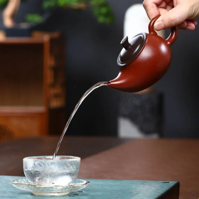 Red teapot pouring tea into a clear glass cup on a wooden table.

