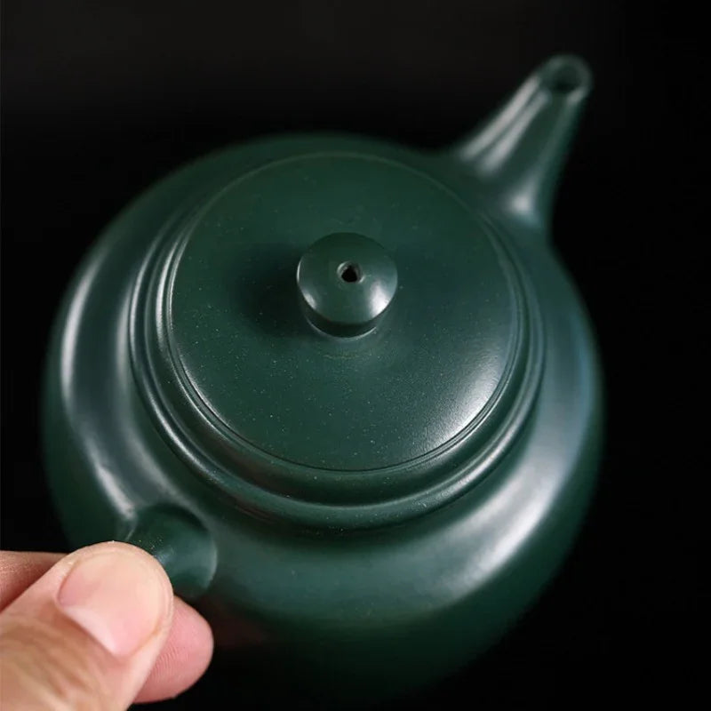 Green teapot held by a hand against a black background

