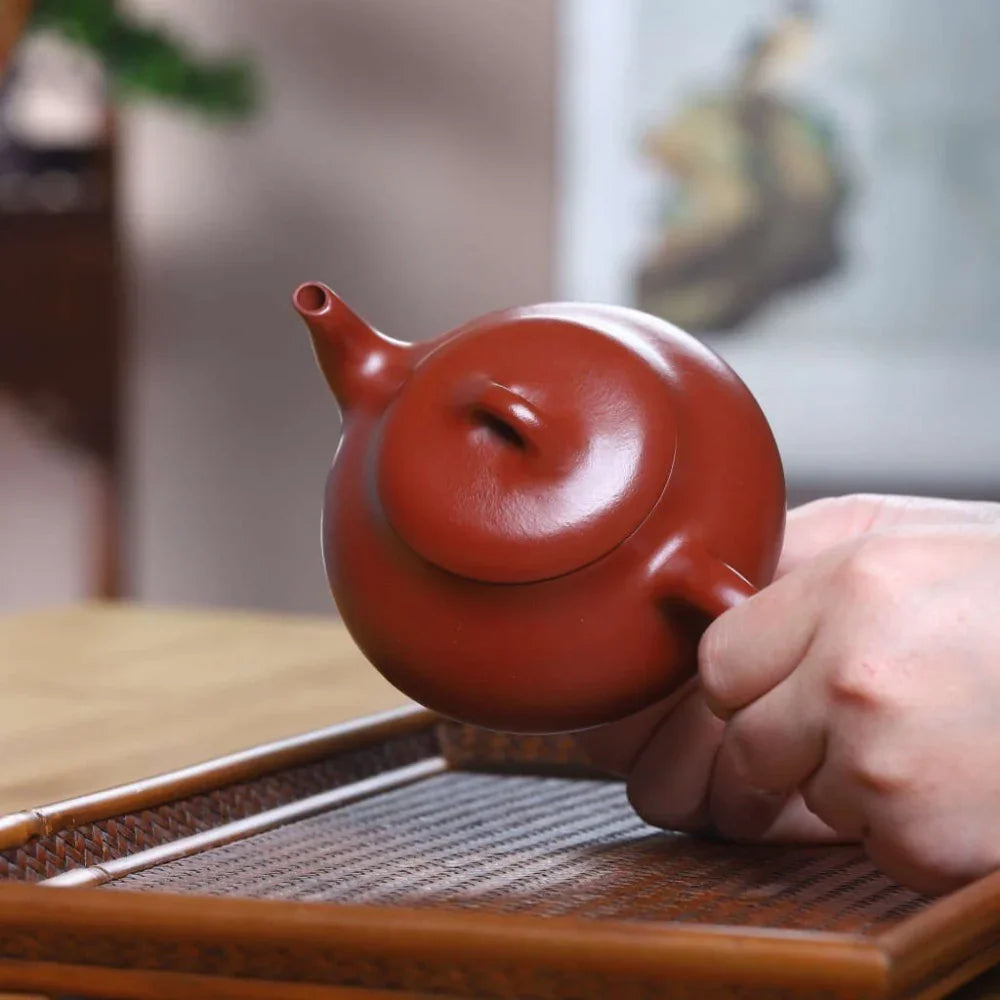 Red ceramic teapot held by a hand on a wooden surface with a blurred background

