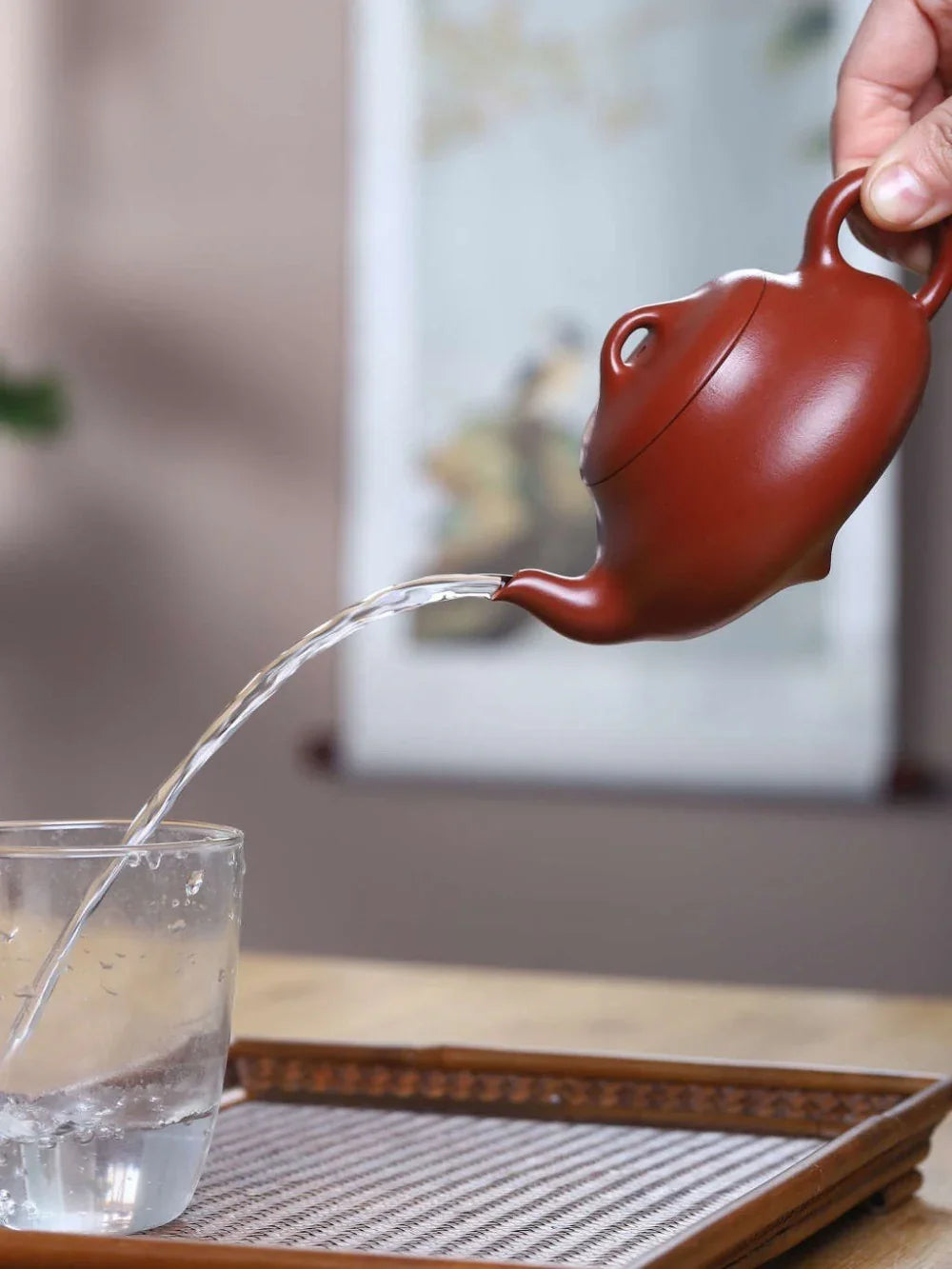Red ceramic teapot pouring water into a glass on a wooden tray.

