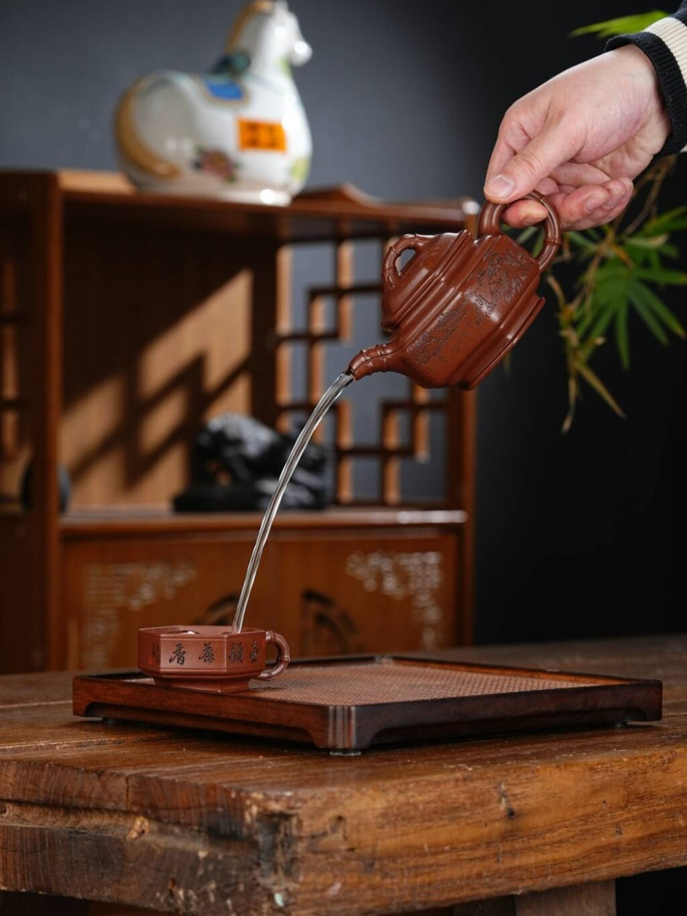 Person pouring tea from a traditional teapot into a cup on a wooden table.

