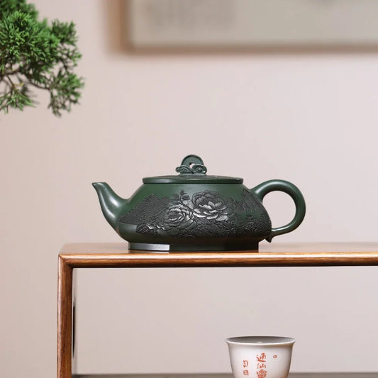 Green ceramic teapot with floral design on a wooden shelf, accompanied by a white cup with red text.

