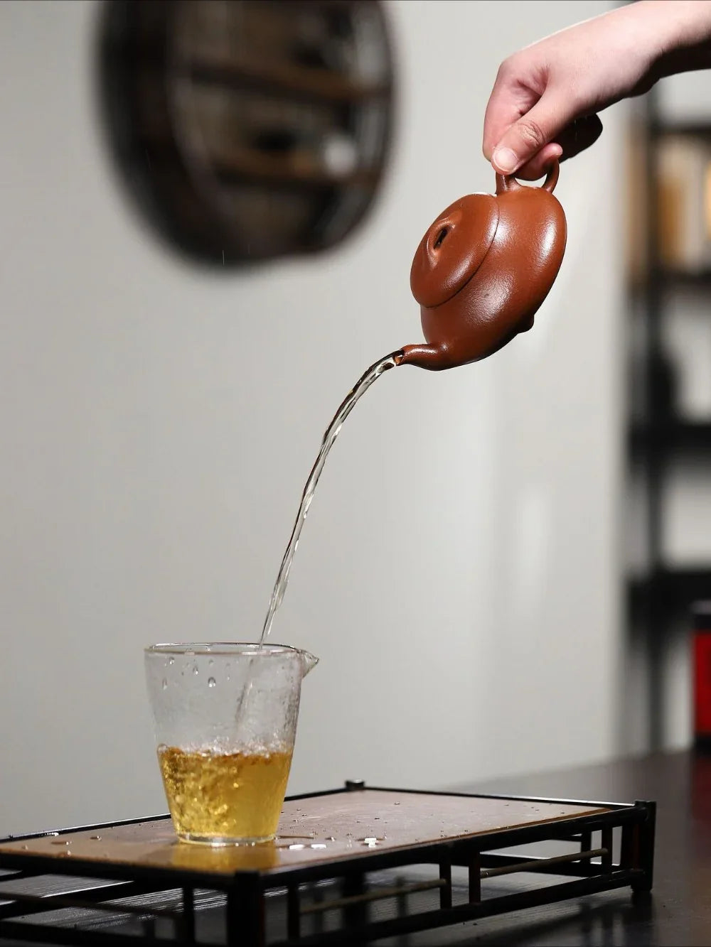 Person pouring tea from a brown teapot into a clear glass cup on a wooden surface.


