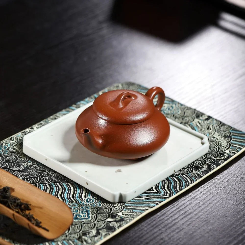 Red ceramic teapot on a white coaster with a wooden tea scoop on a patterned mat.

