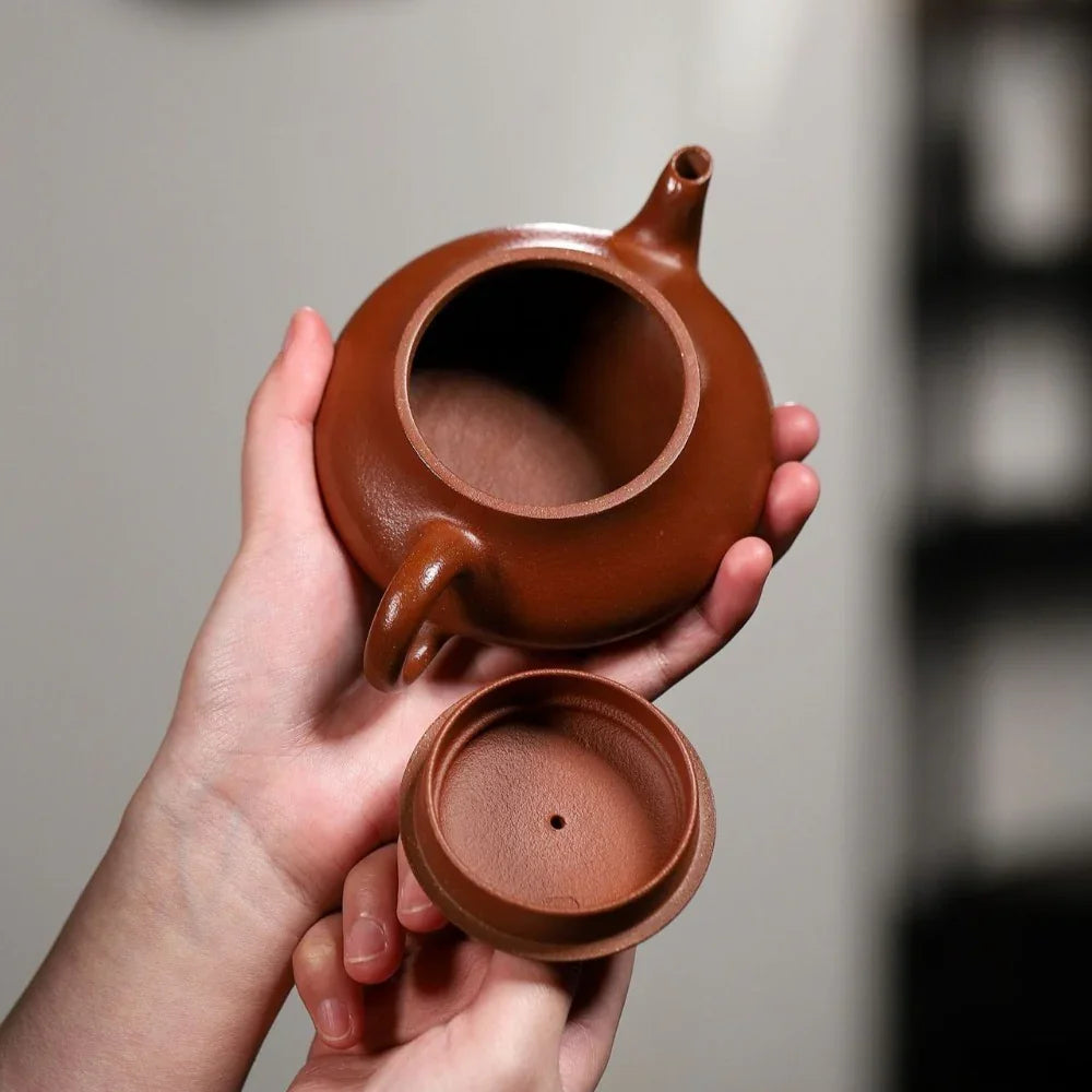 Person holding a brown ceramic teapot and lid against a neutral background

