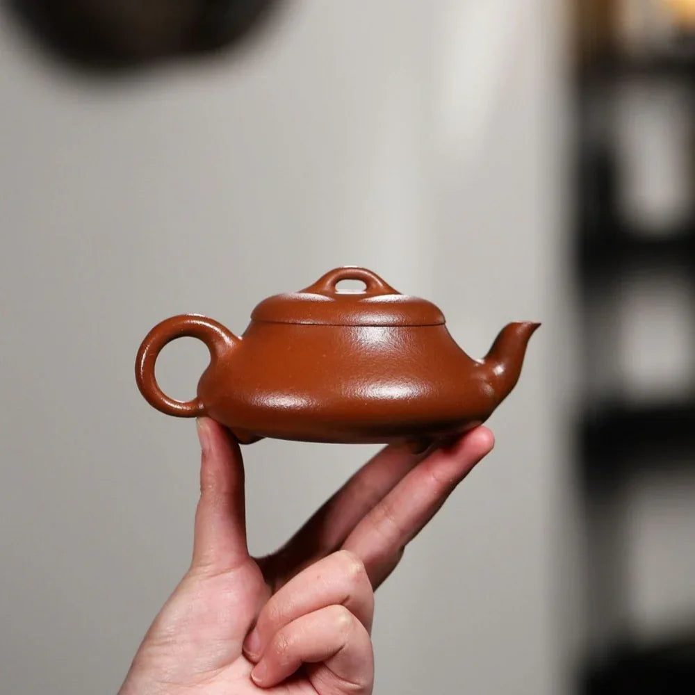 Hand holding a small brown ceramic teapot against a neutral background


