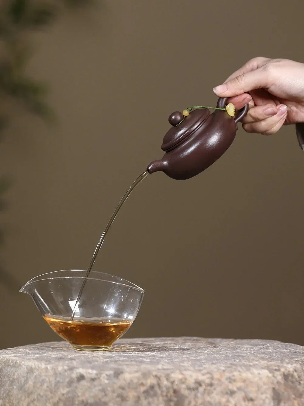 Person pouring tea from a purple teapot into a clear glass cup on a stone surface.

