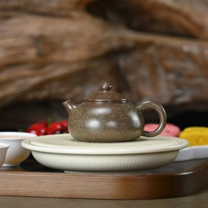 A small speckled brown ceramic teapot sits on a round cream-colored saucer, placed on a wooden tray with a blurred rustic wood background and tea accessories around it.

