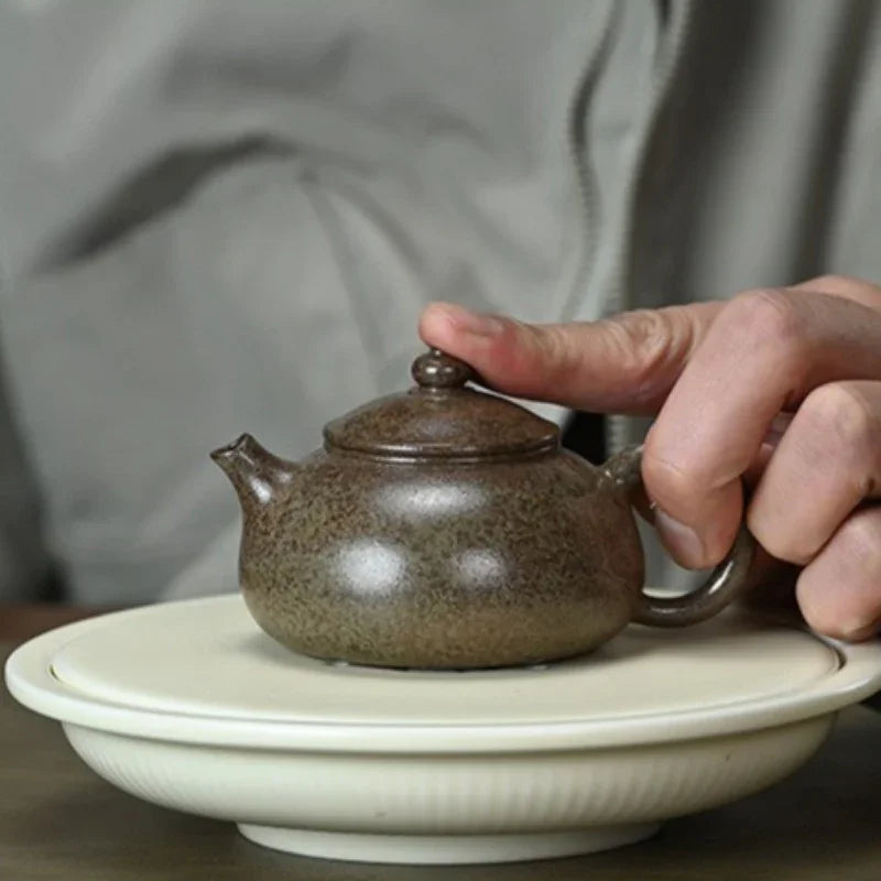 A close-up of a hand holding the handle of a small speckled brown ceramic teapot over a white saucer, with a blurred person in a gray jacket in the background.

