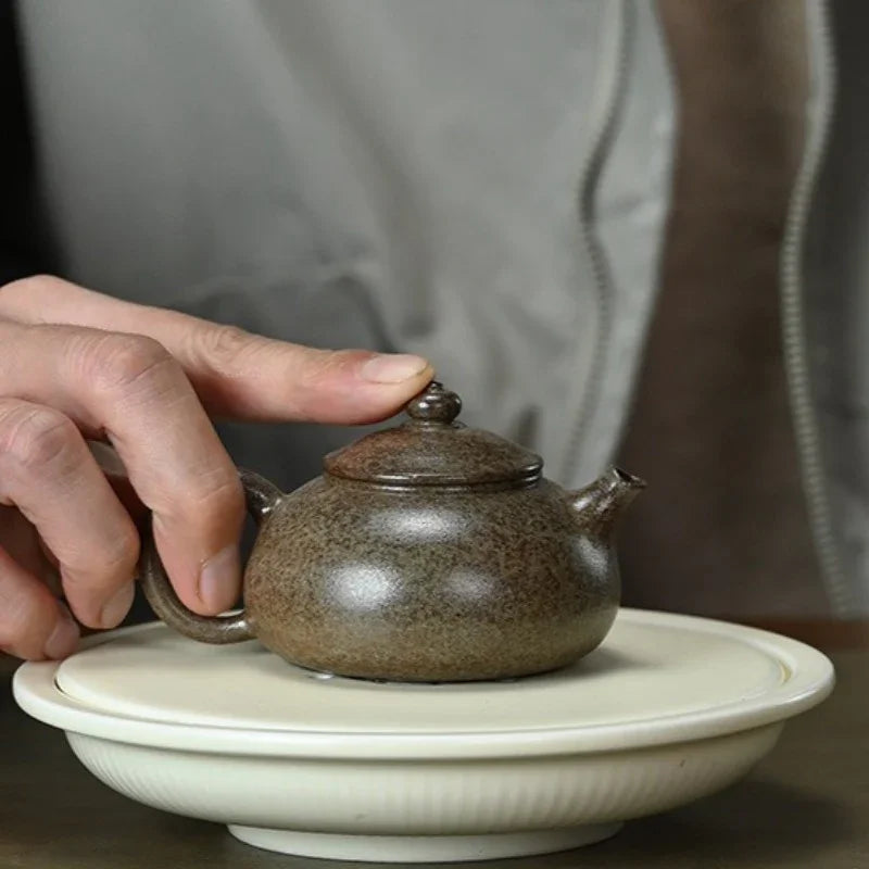 A small speckled brown ceramic teapot with a lid is resting on a white saucer, with a person's hand holding the handle and a gray jacket blurred in the background.

