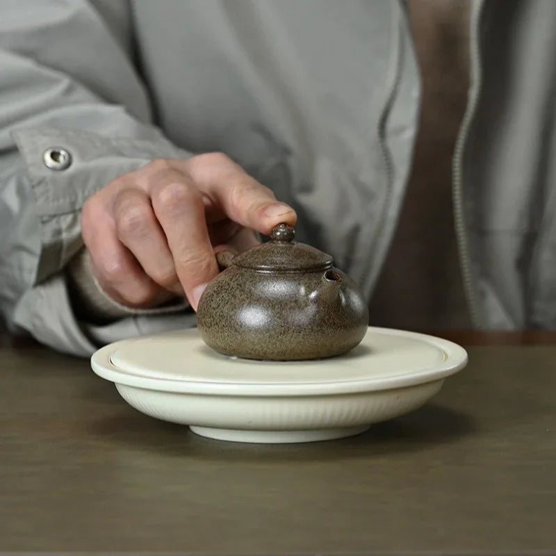 A person in a gray jacket holds the lid of a small speckled brown ceramic teapot resting on a white saucer over a wooden table.

