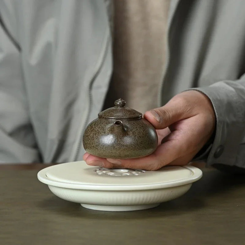 A person's hand holding a small speckled brown ceramic teapot over a white lidded teapot stand on a dark wooden table.

