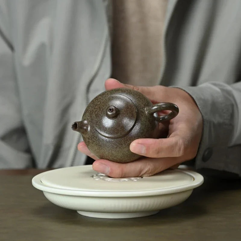 A person's hands holding a small speckled dark brown ceramic teapot over a white ceramic tea tray, with a blurred gray jacket in the background.

